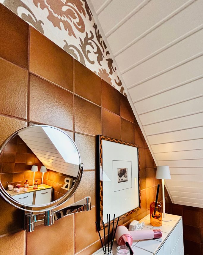 Wand mit braunen Fliesen, dekorativem Muster und Spiegel, dazu ein Bild und Lampe.