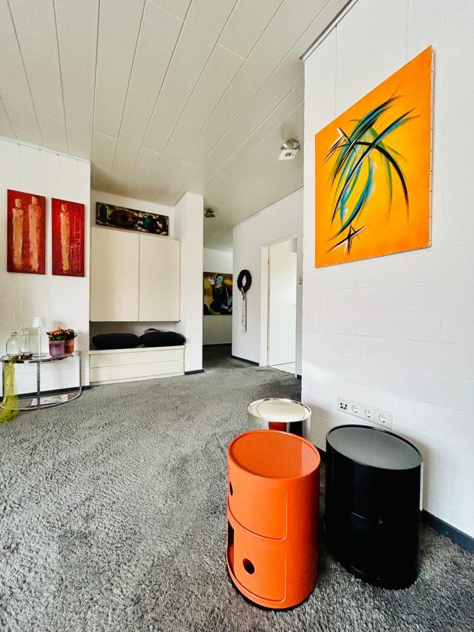 Modernes Zimmer mit orangefarbenem Kunstwerk, farbigen Hockern und minimalistischer Einrichtung.