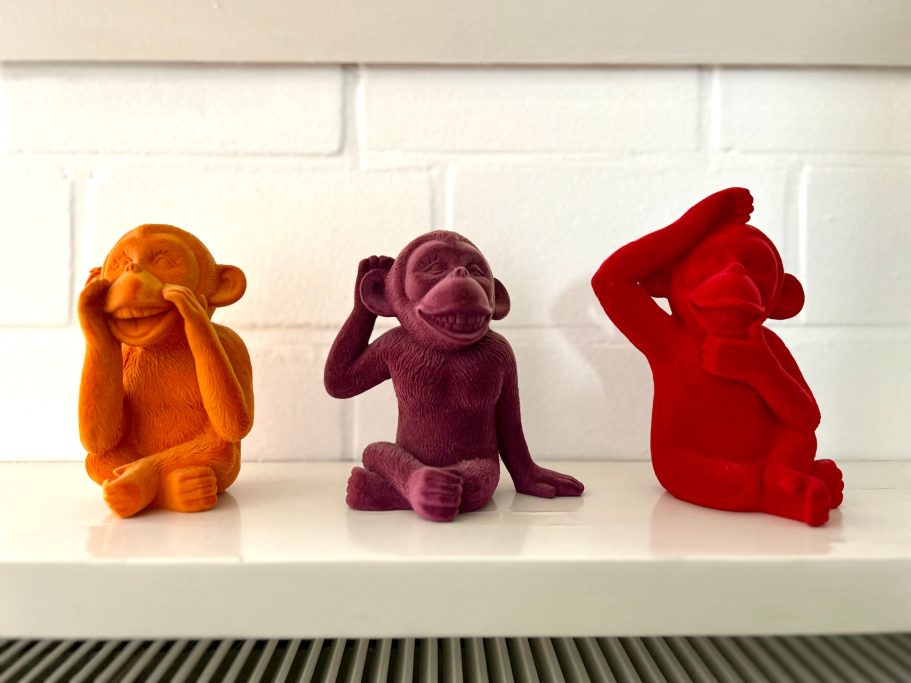Drei bunte Affenskulpturen in Orange, Lila und Rot, sitzen auf einer weißen Fläche.