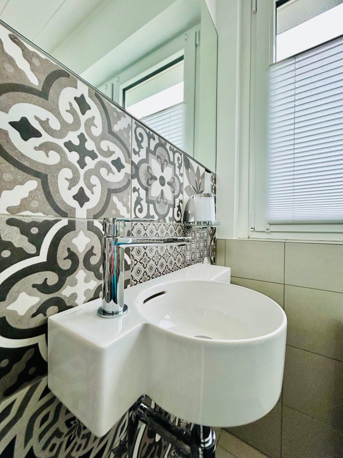 Weiße Wand mit modernem Waschbecken vor kunstvollem, gefliestem Hintergrund.