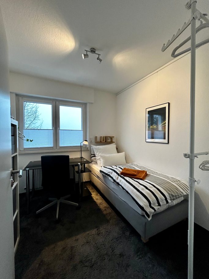 Kleines Zimmer mit Bett, Schreibtisch, Stuhl und Fenster mit Blick nach außen.