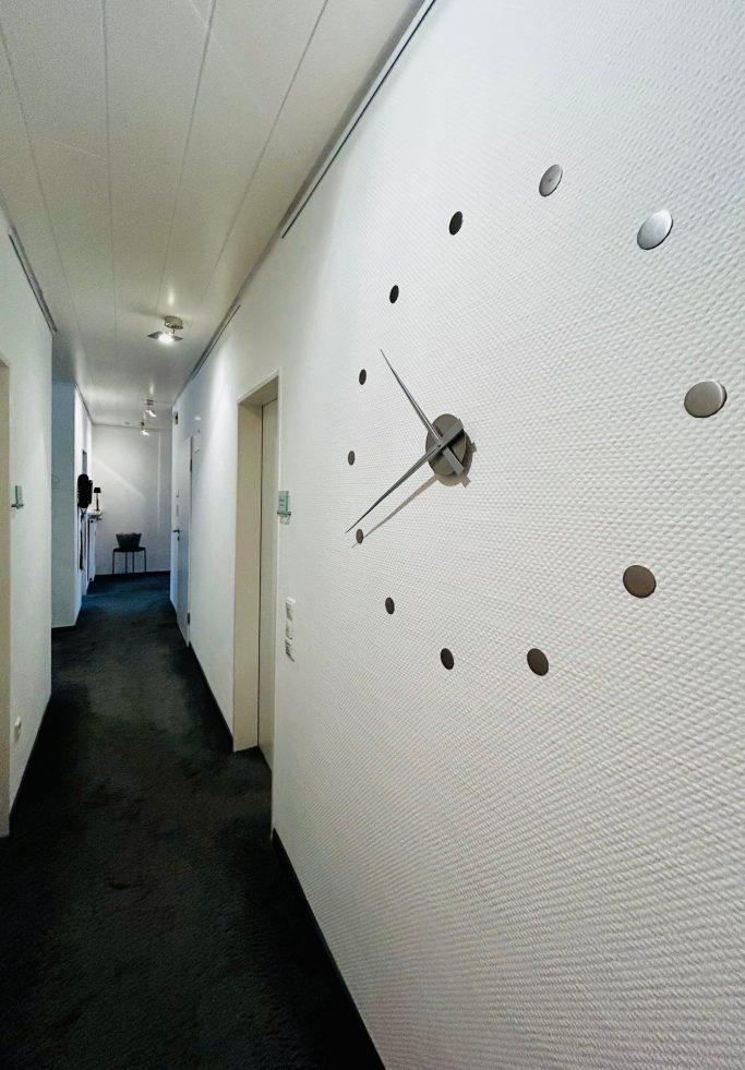 Wand mit einer modernen Uhr und einem langen Flur im Hintergrund.