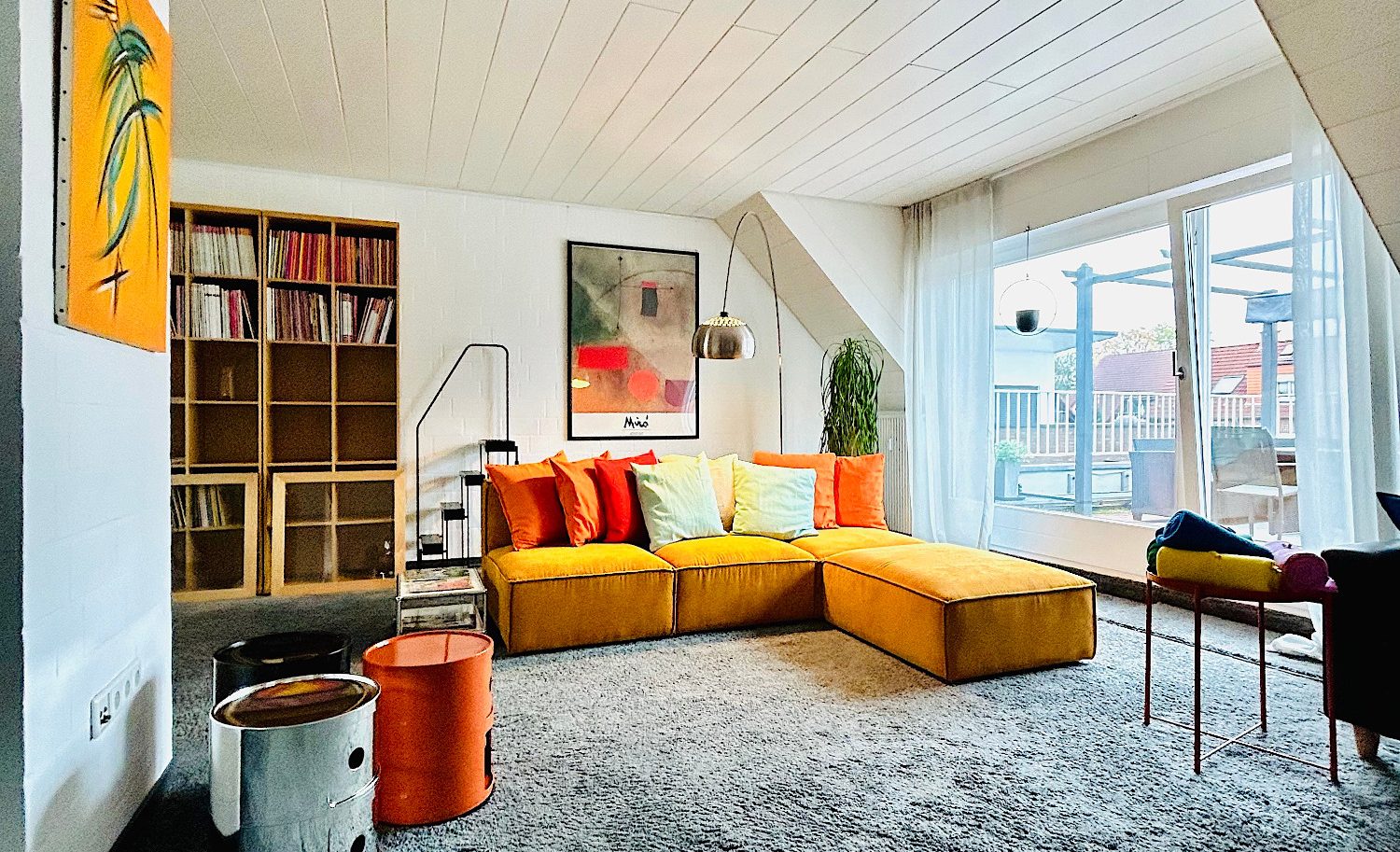 Helle Wohnlandschaft mit bunten Kissen, Bücherregal und großem Fenster.