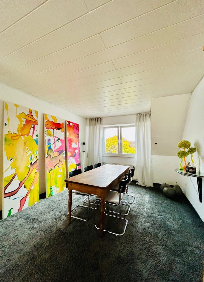 Heller Raum mit bunten Gemälden, einem Holztisch und modernen Möbeln.