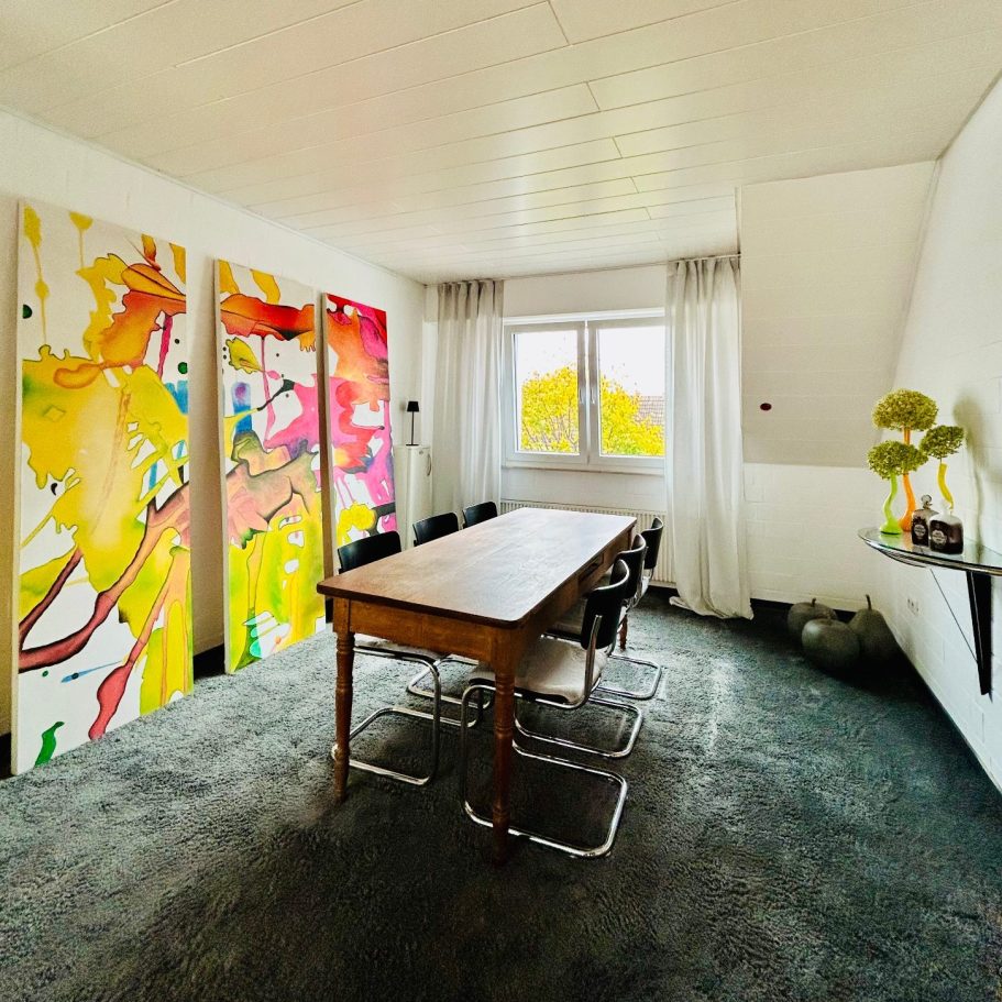 Modernes Zimmer mit einem Holztisch, Stühlen und bunten Wandbildern. Helle Fenster.