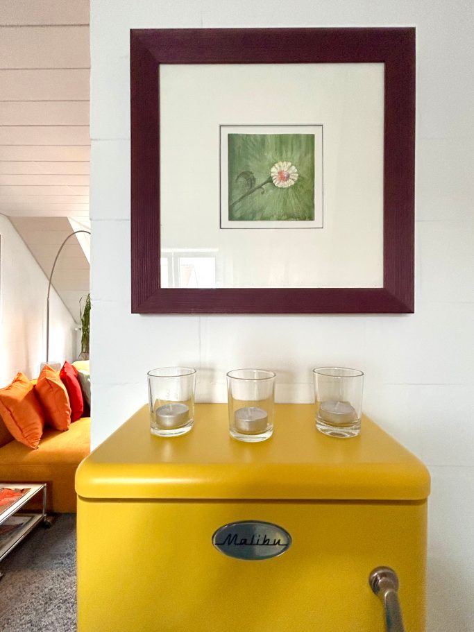 Eine gelbe Kommode mit einem gerahmten Bild und Gläsern darauf, im Hintergrund ein farbenfrohes Sofa.