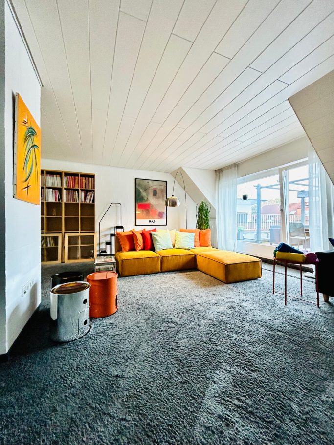 Helles Wohnzimmer mit gelbem Sofa, bunten Kissen und großen Fenstern.